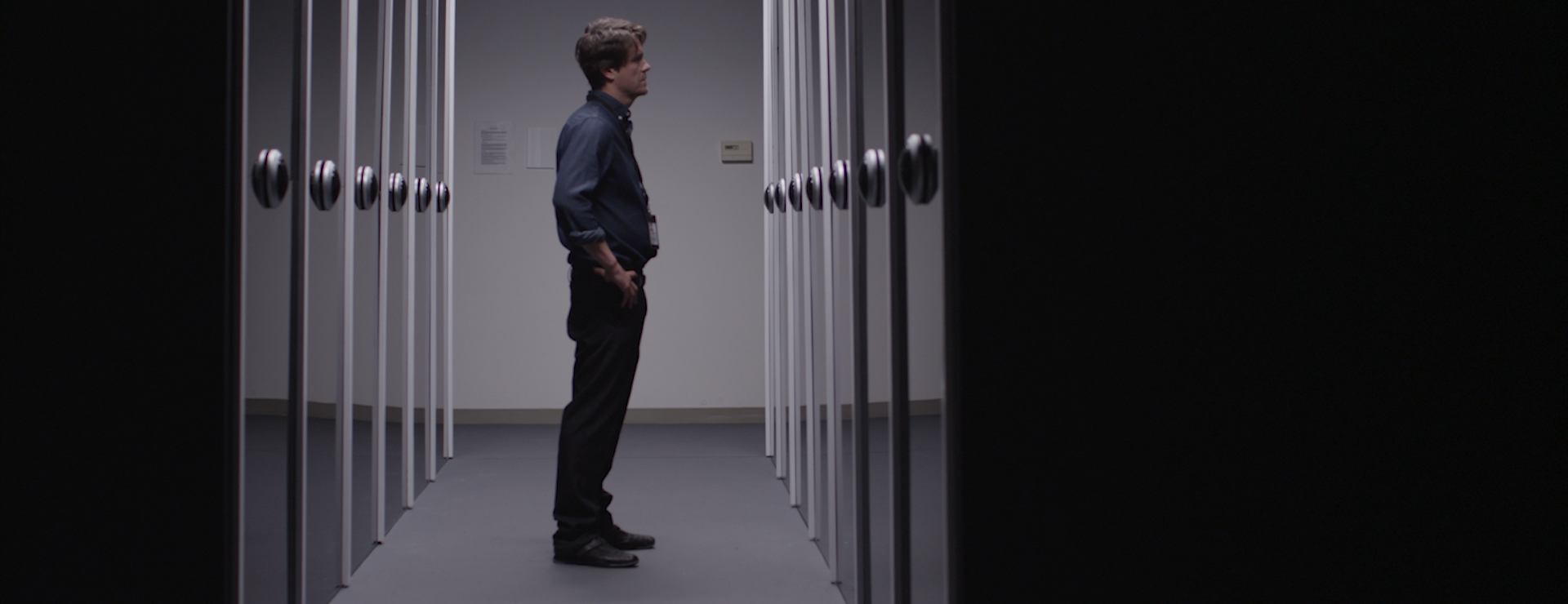 A man stands in a hallway of reflective black glass doors. He is staring into his reflection